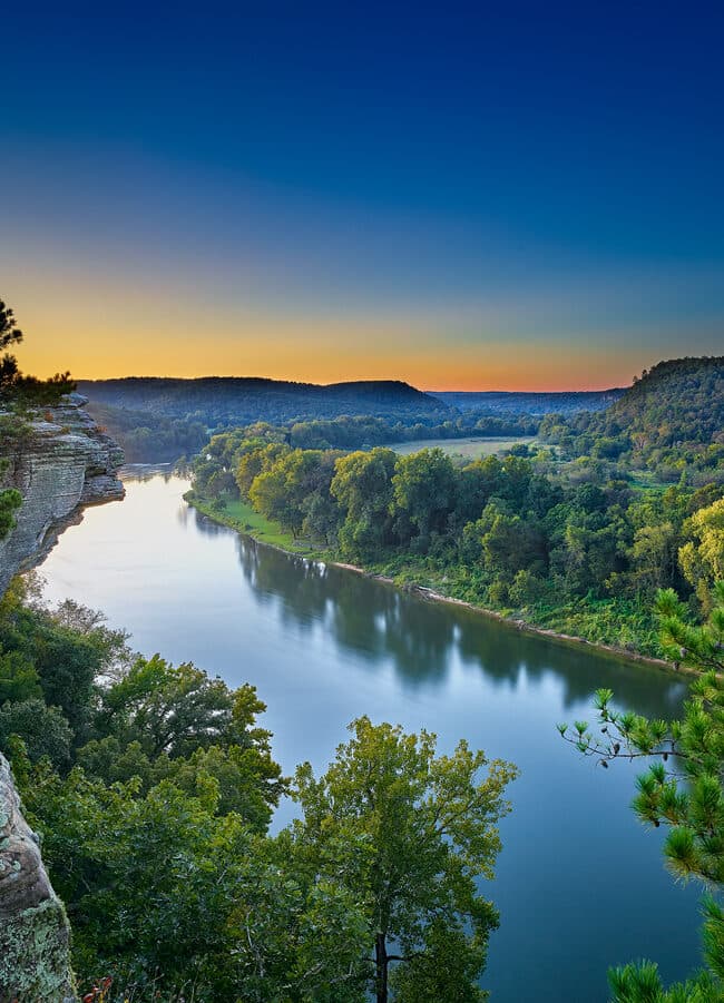 A serene view of a winding river surrounded by lush greenery under a colorful sunrise sky.