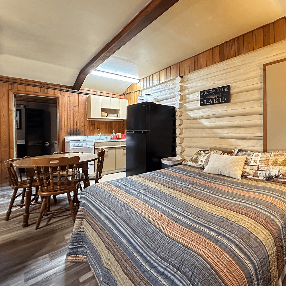 Cozy log cabin interior featuring a dining area, kitchen, and a bed with a colorful quilt.