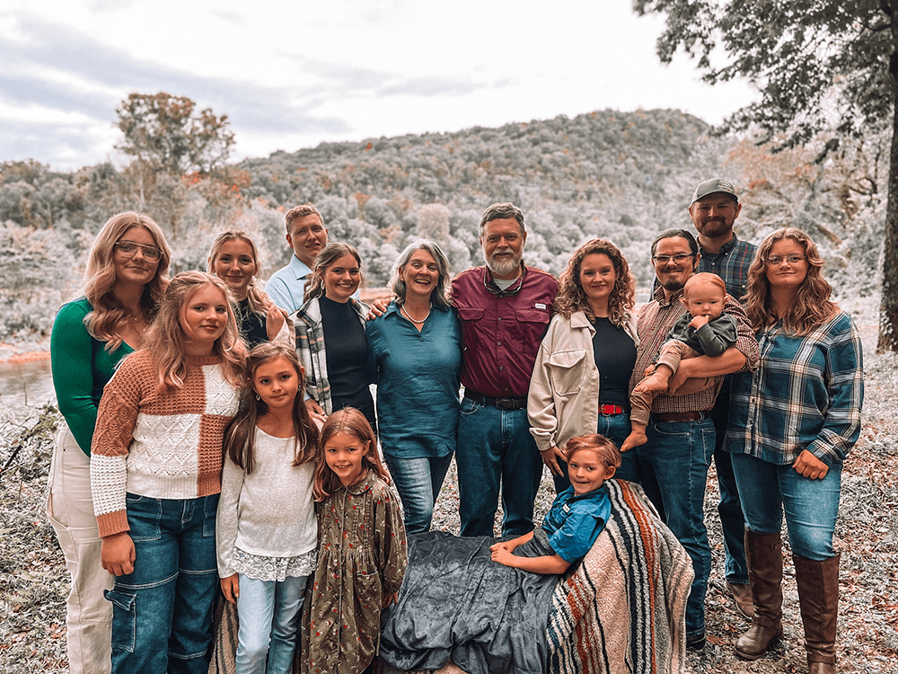 A large group of people poses together outdoors, surrounded by trees and a mountainous backdrop.